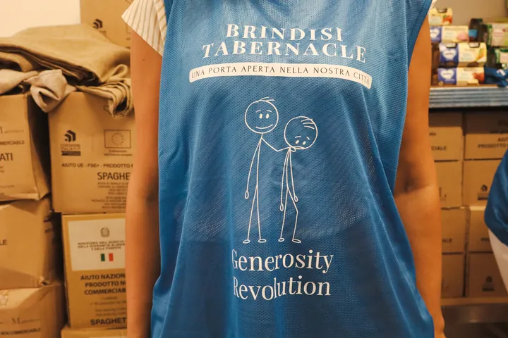 La pettorina blu dei volontari con la scritta Generosity Revolution, tra gli scatoloni del magazzino