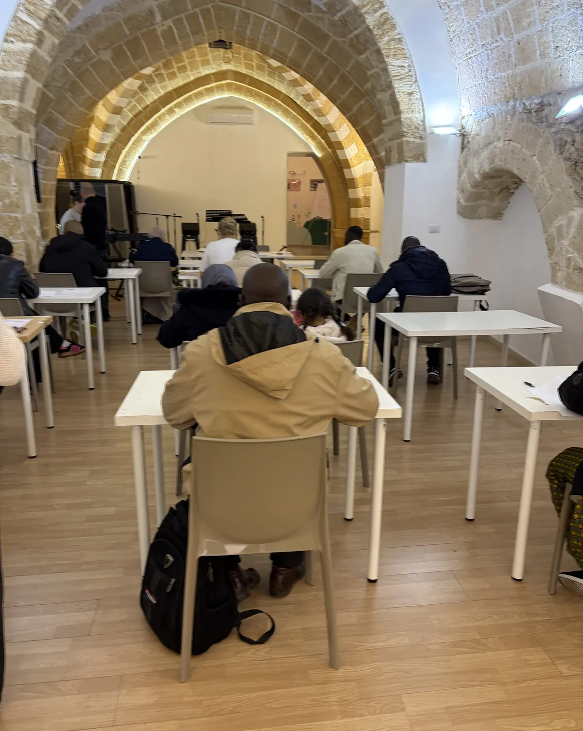 Adulti seduti ai banchi durante una lezione di italiano, nella sala con gli archi in pietra