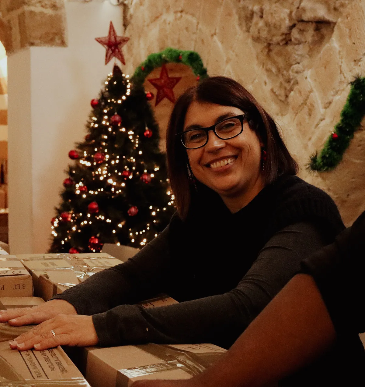 A volunteer smiles leaning on boxes of food ready to go, in a stone-walled room