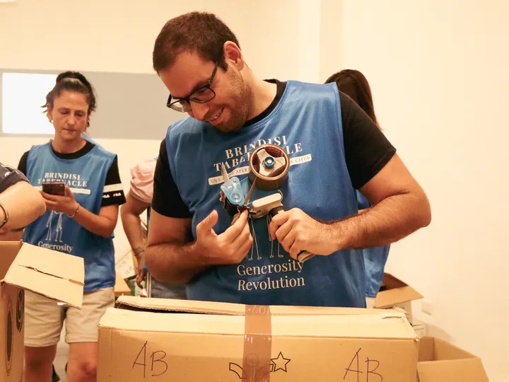 Un volontario con la pettorina blu chiude con il nastro uno scatolone di alimenti al Dream Center